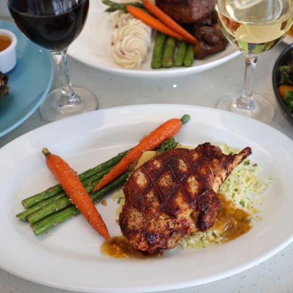 Grilled chicken with rice, asparagus, and carrots, with wine glasses in the background at Lulu California Bistro.