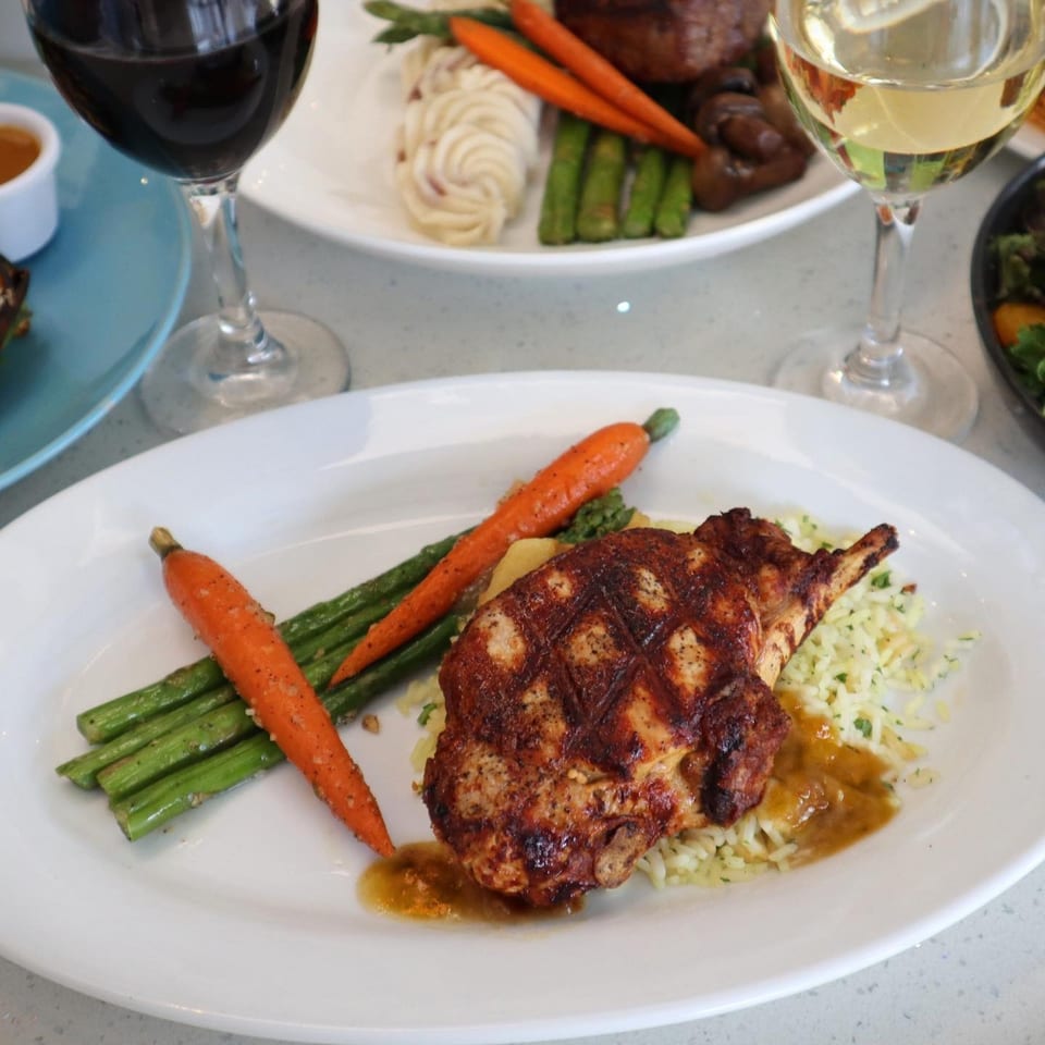 Grilled chicken with rice, asparagus, and carrots, with wine glasses in the background at Lulu California Bistro.