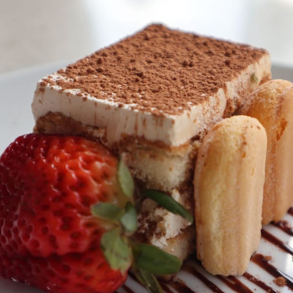 Tiramisu with cocoa powder, ladyfingers, a fresh strawberry, and chocolate drizzle on a white plate.
