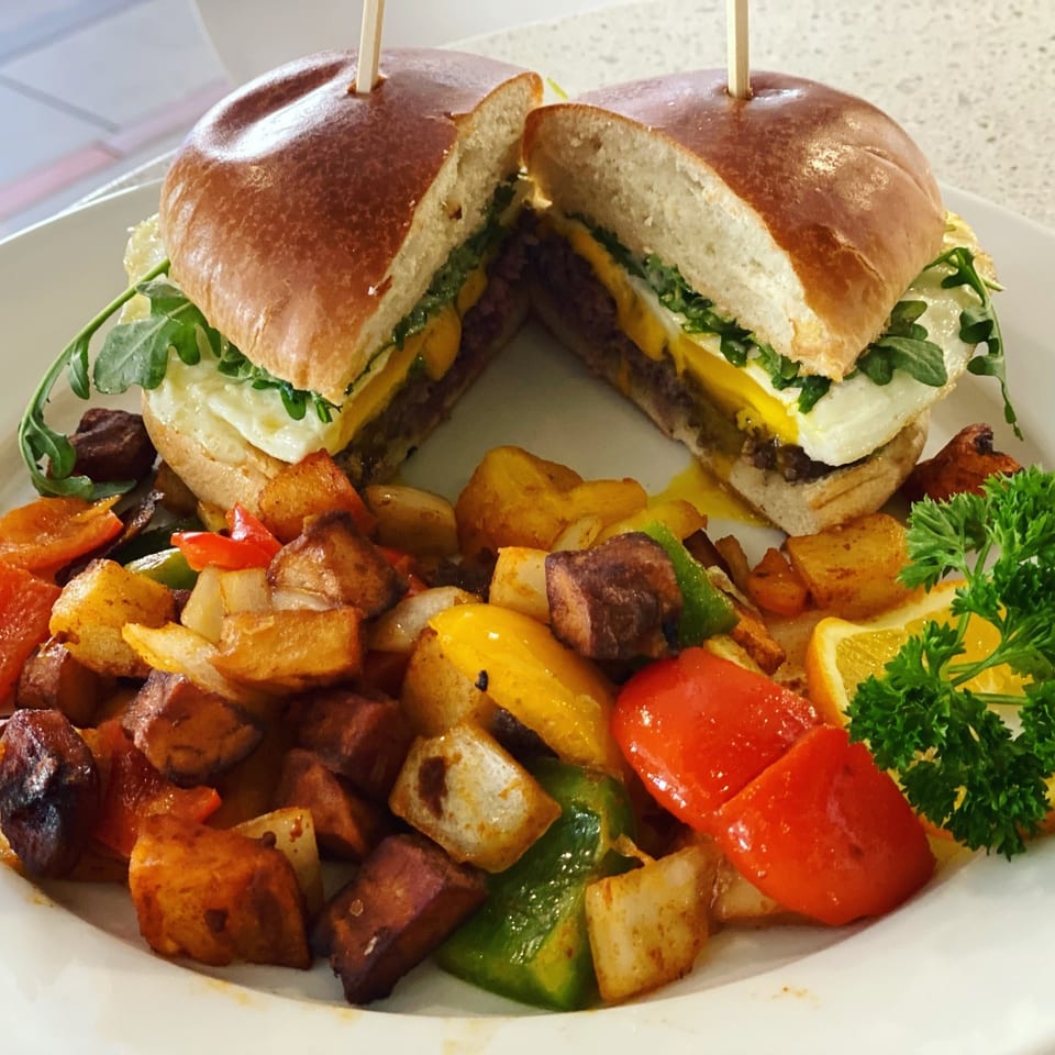 A breakfast burger with egg, cheese, and arugula, served with roasted potatoes and bell peppers.