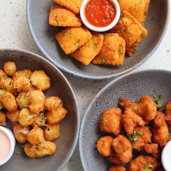 Three bowls of golden-fried appetizers: ravioli with marinara, cheese curds with sauce, and breaded cauliflower.