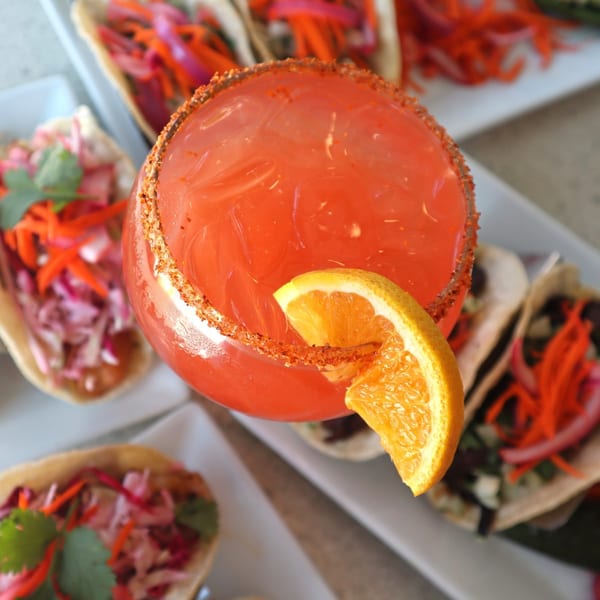 Overhead view of a vibrant red cocktail with an orange slice and chili-lime rim, surrounded by tacos.