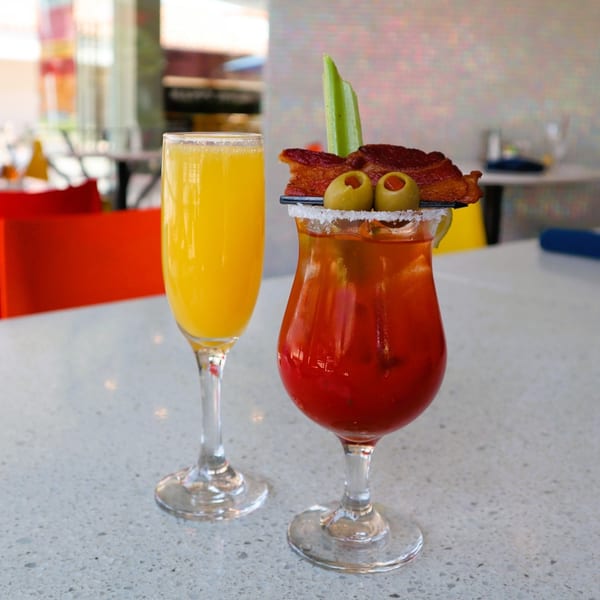A mimosa and a Bloody Mary garnished with bacon, olives, and celery sit on a table.